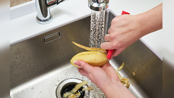 A person peels a potato with a red peeler under running water in a stainless steel kitchen sink, following plumbing tips from professional plumbers to prevent clogs.