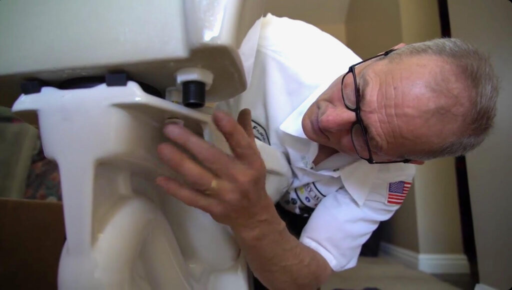 A plumber wearing glasses and a uniform inspects the underside of a toilet tank inside a home, searching for toilet problems that may need quick fixes.
