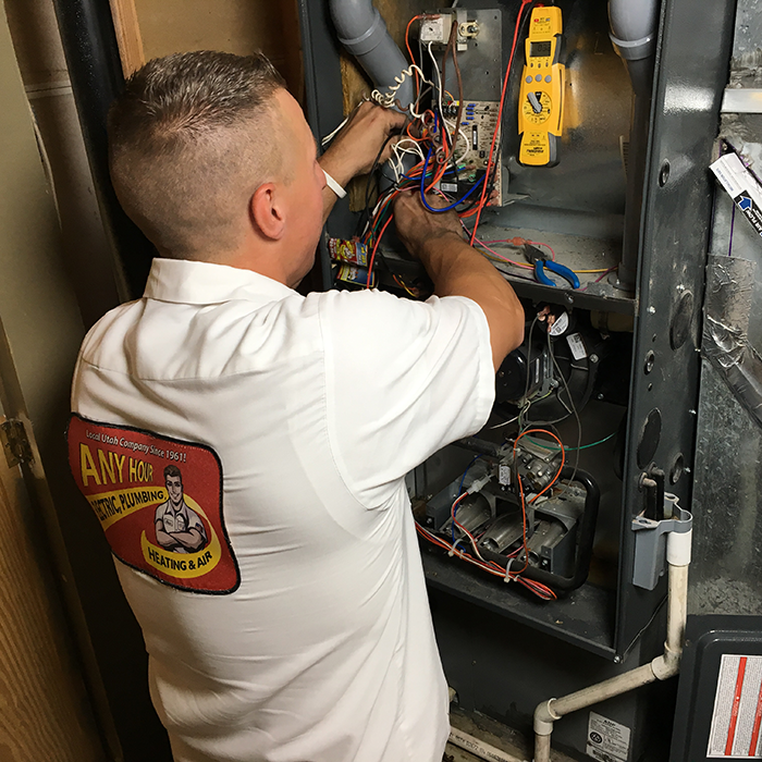 A technician in a white shirt labeled "ANY HOUR ELECTRIC PLUMBING HEATING & AIR" performs furnace maintenance, carefully working on the wiring inside a furnace.