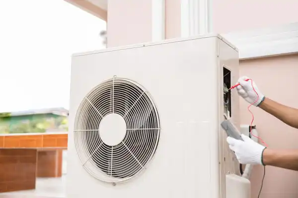 A technician wearing white gloves tests an outdoor air conditioning unit with a multimeter during heat pump maintenance in Salt Lake City.