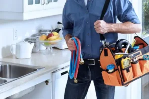 A person in a blue shirt stands in a Salt Lake City kitchen holding hoses and a tool bag filled with kitchen plumbing repair tools near the sink.