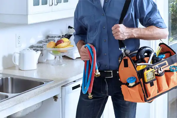 A person in a blue shirt stands in a Salt Lake City kitchen holding hoses and a tool bag filled with kitchen plumbing repair tools near the sink.