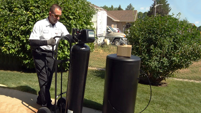 A technician in uniform moves a black water softener tank on a dolly next to another large black tank and a shrub, installing water conditioners to address hard water in a residential yard.