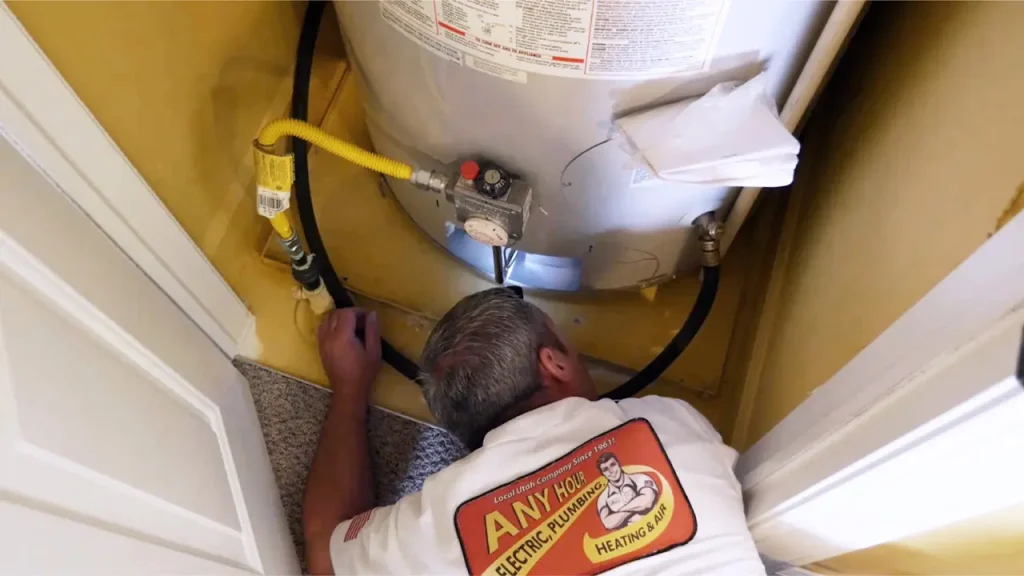 A plumber in a white shirt is inspecting pipes beneath a water heater in a small, yellow-walled utility closet, ensuring optimal water heating performance.