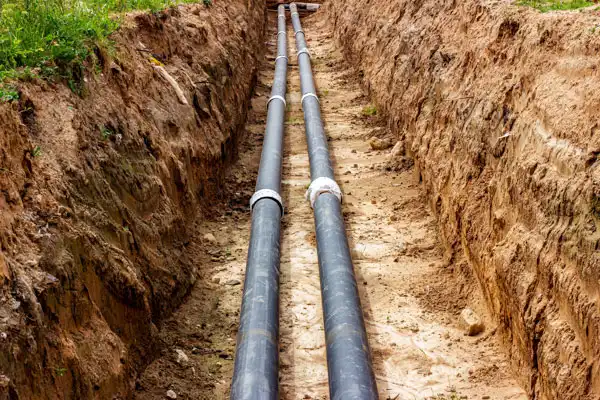 Two black plastic pipes, part of a sewer line installation in Salt Lake City, are laid parallel in a narrow, excavated trench surrounded by dirt and grass at the edges.