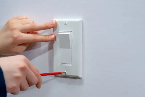 A person uses a screwdriver to remove a screw from the bottom of a white electrical switch plate on a wall, preparing for outlets service in Salt Lake City.