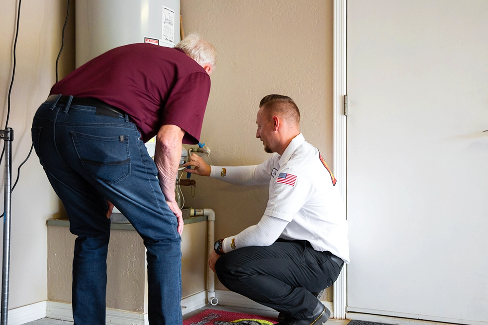 A technician kneels and explains a water heater to an older man standing beside him in a residential utility room.