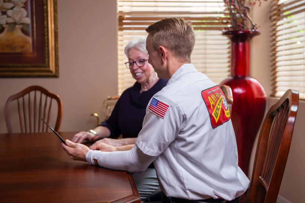 %Imagetitle% | Any Hour Services A technician in uniform shows a tablet to an older woman at a dining table in a well-lit room, discussing available financing options.