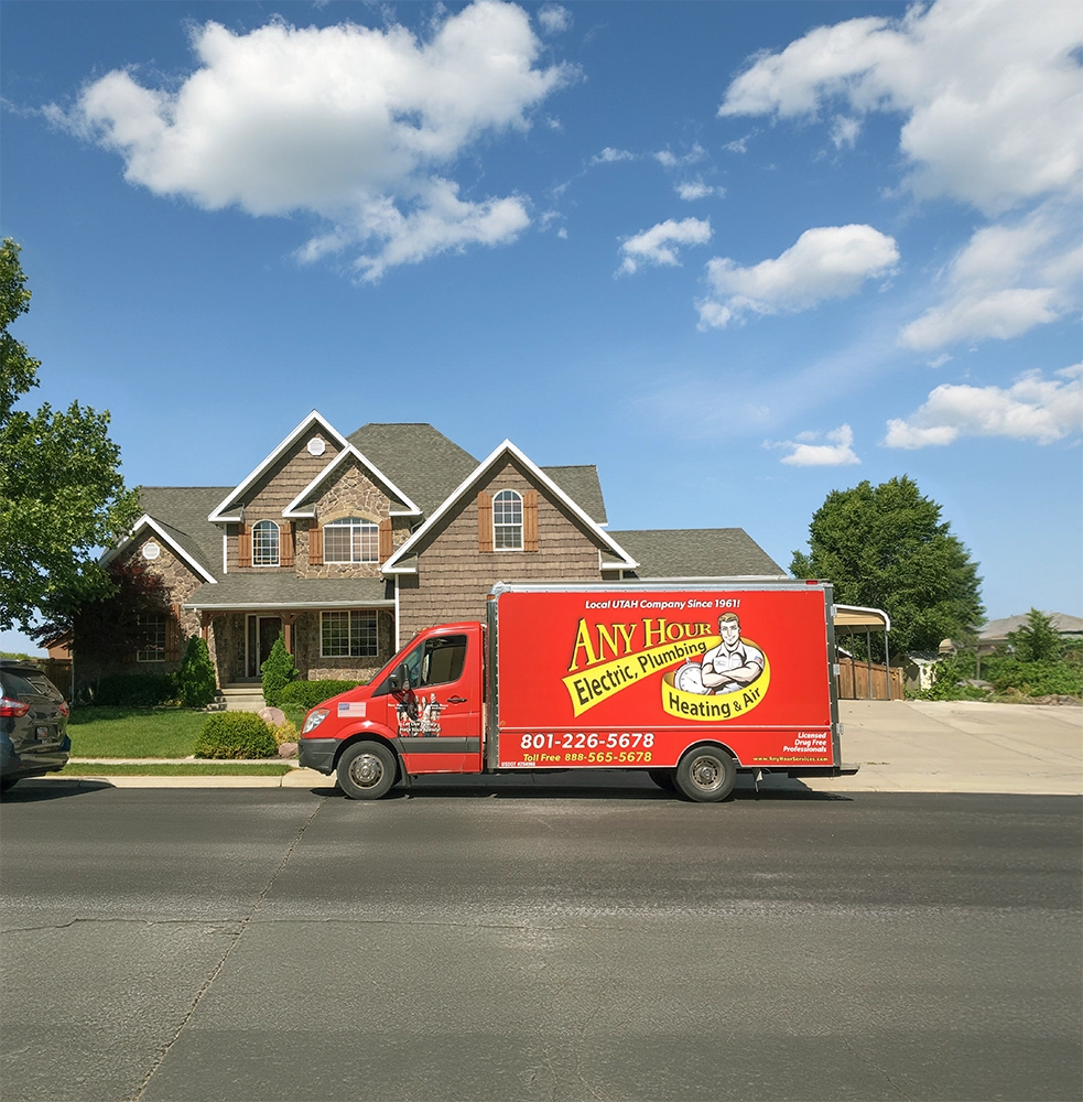 A red Any Hour Electric, Plumbing, Heating & Air service van is parked on the street in front of a large suburban house on a sunny day.