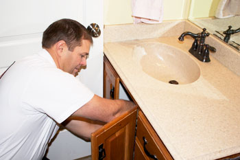 A man kneels and works under a bathroom sink, reaching into the cabinet below the countertop to address plumbing issues.