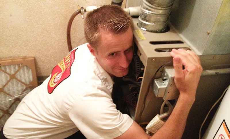 A technician from the Any Hour Services team in uniform is inspecting and embracing a furnace in a utility room, with various wires and components visible.