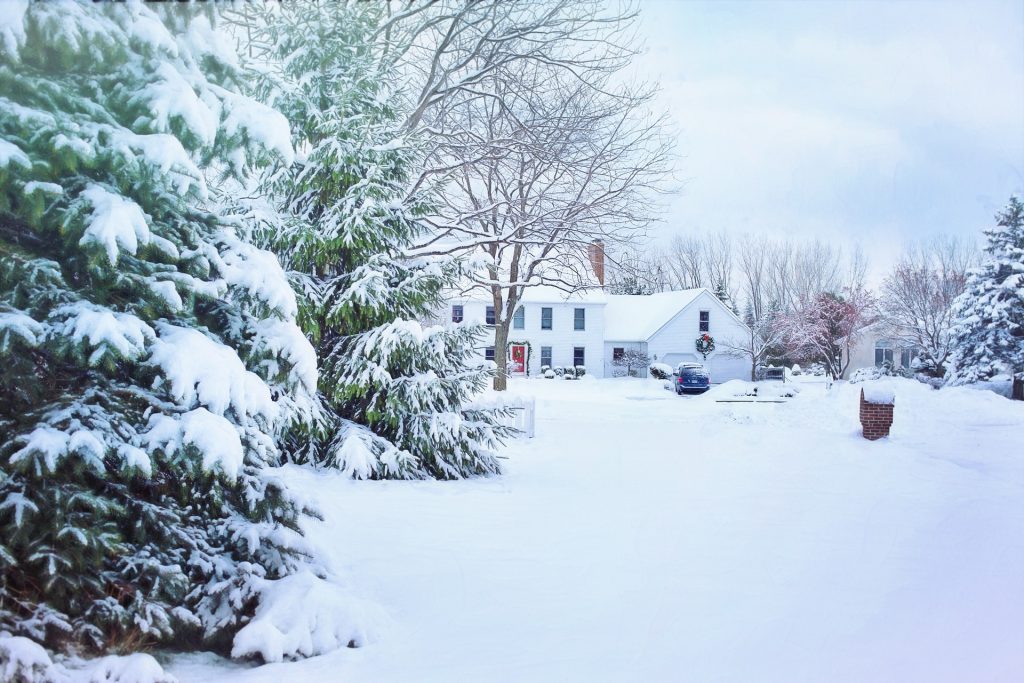 A white house with black shutters and a wreath on the door sits behind snow-covered trees and a driveway, showcasing the charm of winter home preparation after a recent snowfall.