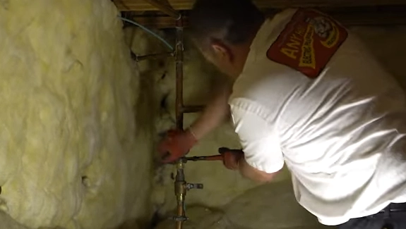 A person wearing gloves and a white shirt uses a wrench to work on copper pipes near insulation in a basement, ensuring optimal water pressure for the system.