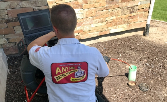 A technician wearing an "Any Hour Electric, Plumbing, Heating & Air" shirt inspects outdoor plumbing with a camera and monitor next to a building, checking for sewer signs that could indicate the need for sewer repair.