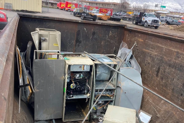 Large metal appliances and HVAC units from equipment disposal are discarded inside an industrial dumpster in a parking lot, with vehicles and buildings visible in the background.