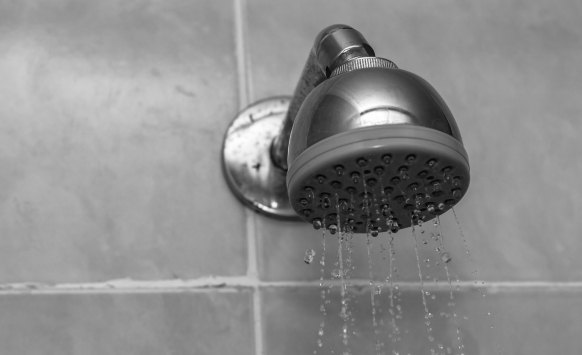 A close-up of a showerhead with water lightly spraying out, mounted on a tiled wall, showcasing the plumbing and gentle water pressure.