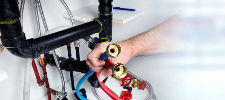 A person holds two flexible water hoses near plumbing pipes and valves under a sink, preparing for plumbing installation or repair in Salt Lake City.