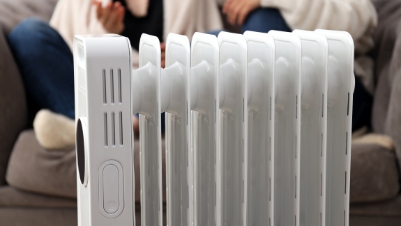 A white oil-filled space heater is placed in front of a sofa, with two people sitting in the background, partially visible.