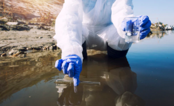 Person in protective gear collects a water sample from a river or lake using a vial and container, likely for environmental testing of various water contamination categories.