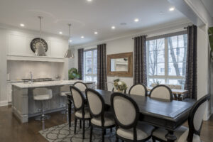 %Imagetitle% | Any Hour Services Modern kitchen and dining area with white cabinets, a dark dining table, upholstered chairs, bar stools, pendant lights, and large windows with patterned curtains.