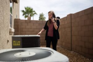 %Imagetitle% | Any Hour Services A woman stands outside next to two air conditioning units, talking on her phone and inspecting one of the units near a brick wall.