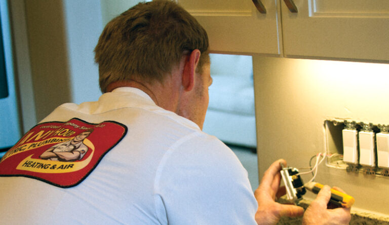 A technician in a white shirt is working on an electrical outlet, using tools to adjust wiring below a row of light switches—demonstrating basic electrical skills for DIY electrical upgrades.