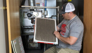 %Title% | Any Hour Services A man examines a dirty air filter in front of an open furnace unit inside a closet.
