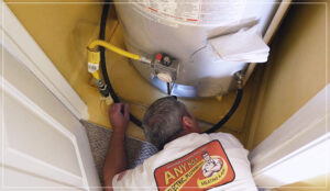 %Title% | Any Hour Services A plumber in a branded shirt inspects the connections at the base of a water heater inside a small utility closet, checking for signs of water heater failure.