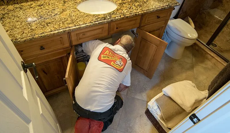 %Title% | Any Hour Services A plumber kneels on a red pad while working under a bathroom sink, with cabinet doors open and tools nearby—showcasing the plumbing knowledge professional plumbers bring to every job.