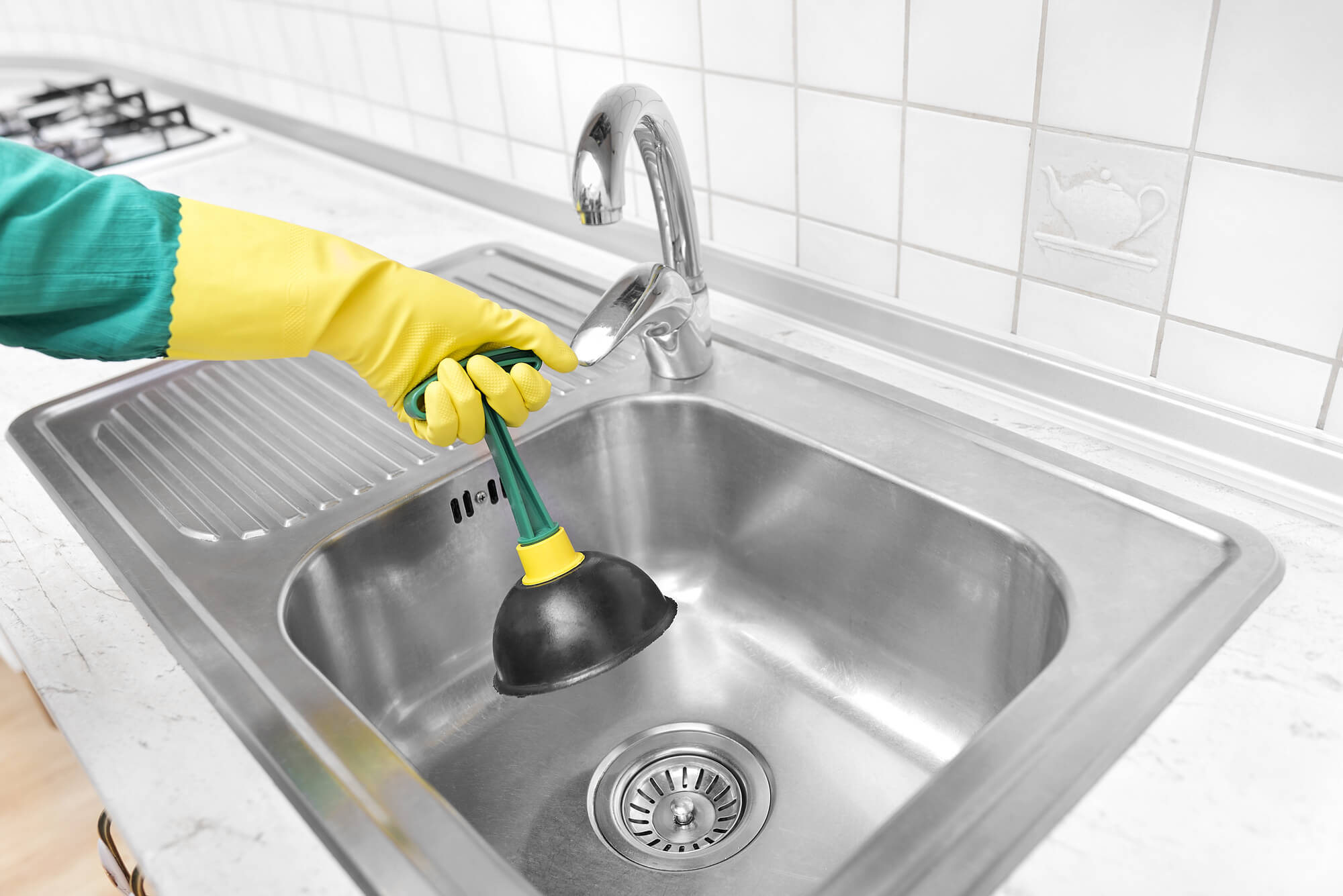 Person wearing yellow rubber gloves uses a plunger to unclog a stainless steel kitchen sink under a chrome faucet, choosing manual effort over chemical drain cleaners.