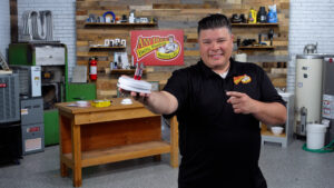 A man in a black polo shirt stands indoors, smiling and pointing at a white and red device he is holding—a chirping smoke detector— with a workbench and tools in the background.