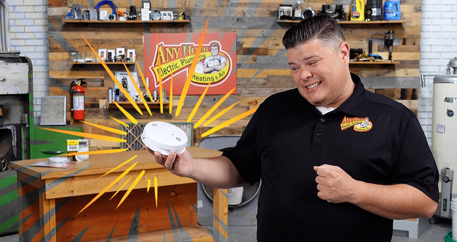 %Imagetitle% | Any Hour Services A man in a black polo shirt holds a smoke detector with animated yellow rays around it, standing in a workshop filled with tools—ready to share tips on smoke detector troubleshooting and how to fix smoke detector issues.
