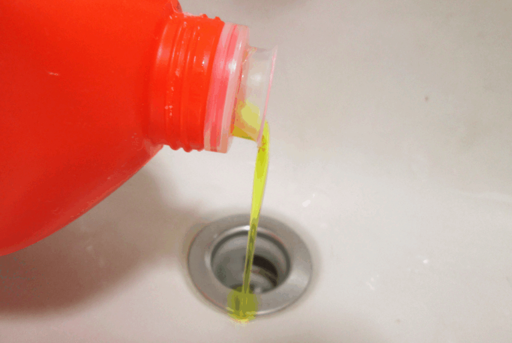 A red bottle pours yellow-green liquid down a metal sink drain, highlighting potential chemical drain cleaner problems.