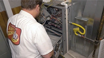 A technician wearing a logo shirt inspects and repairs the interior components of a residential furnace during routine furnace maintenance in a utility room.