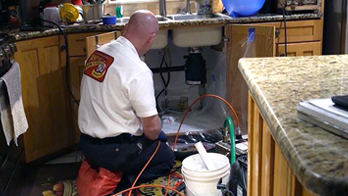 A plumber works under a kitchen sink, using tools and equipment to fix plumbing issues with cabinet doors open and supplies nearby, preparing for advanced techniques like pipe jetting to clear stubborn clogs.
