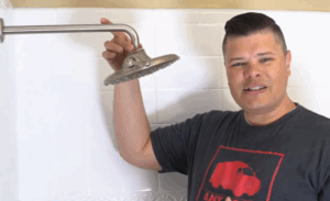 %Title% | Any Hour Services A person stands in a shower, holding a large round shower head attached to a metal arm against a tiled wall, ready to remove hard water buildup.