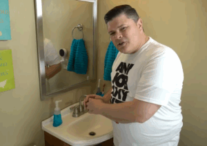 %Title% | Any Hour Services A man in a white t-shirt stands at a bathroom sink, turning on the faucet, with blue towels and soap dispenser visible—possibly about to fix a slow bathroom sink drain.