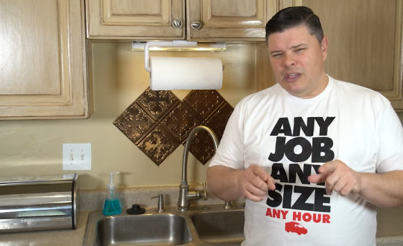 A man wearing a white "ANY JOB ANY SIZE ANY HOUR" shirt stands beside a kitchen sink, gesturing with his hands—demonstrating how to clean sink drain. Kitchen cabinets, soap, and a roll of paper towels are visible.