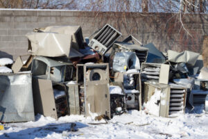 A pile of damaged and dismantled HVAC units and metal appliances sits outdoors on snow near a cinder block wall, serving as a reminder of the importance of HVAC efficiency and proper home ventilation.