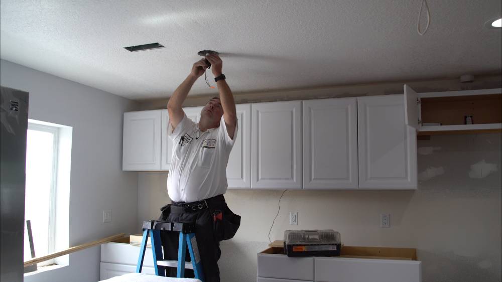 %Title% | Any Hour Services An electrician stands on a ladder installing a ceiling light fixture in a kitchen with white cabinets, ensuring the Arizona home’s lighting complements its HVAC system.