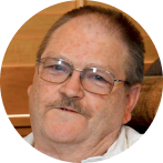 %Focuskw% | Any Hour Services An older man with glasses, a mustache, and thinning hair is seated indoors in front of a wooden background, looking slightly off-camera.