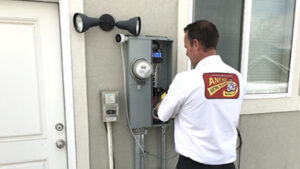 A technician in a white uniform inspects an outdoor electrical panel mounted on a wall next to a door and a security light, ensuring home safety with careful attention to electrical safety.