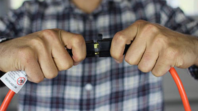 A person in a plaid shirt is disconnecting two black electrical plugs attached to an orange cord, demonstrating good electrical safety by handling the connection with care.