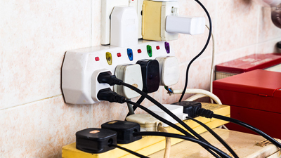 Multiple electrical plugs and adapters are connected to extension sockets and power strips on a wall, creating an overloaded outlet setup—highlighting the importance of electrical safety in your home.