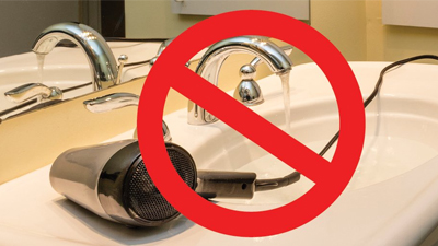 A hair dryer with a cord lies in a bathroom sink with running water, overlaid by a red prohibition symbol—a clear reminder of electrical safety at home.