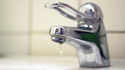 %Imagetitle% | Any Hour Services A single drop of water falls from a chrome bathroom faucet against a white tiled background—a subtle sign of common plumbing issues that may require attention or plumbing repair.