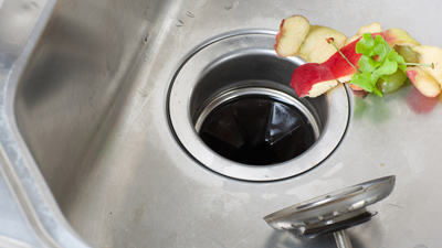 %Imagetitle% | Any Hour Services A stainless steel kitchen sink with food scraps, including apple peels and lettuce, near the drain—a setup that can lead to common plumbing problems requiring plumbing repair. A metal drain stopper sits on the side.
