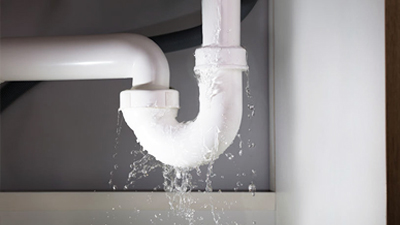 %Imagetitle% | Any Hour Services White PVC pipes under a sink are leaking water at a joint, with water streaming down from the connection—an example of common plumbing issues found in many households.