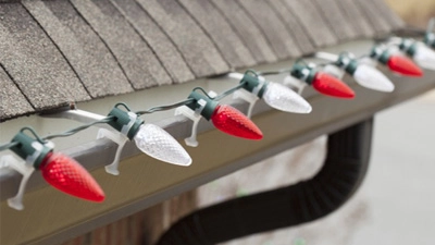 Red and white holiday Christmas lights are clipped along the edge of a house roof above a gutter, powered by an extension cord for Christmas lights.