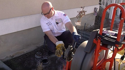 %Title% | Any Hour Services A plumber wearing gloves operates a drain cleaning machine outside a house, clearing a clogged outdoor drain caused by a sewer line issue.